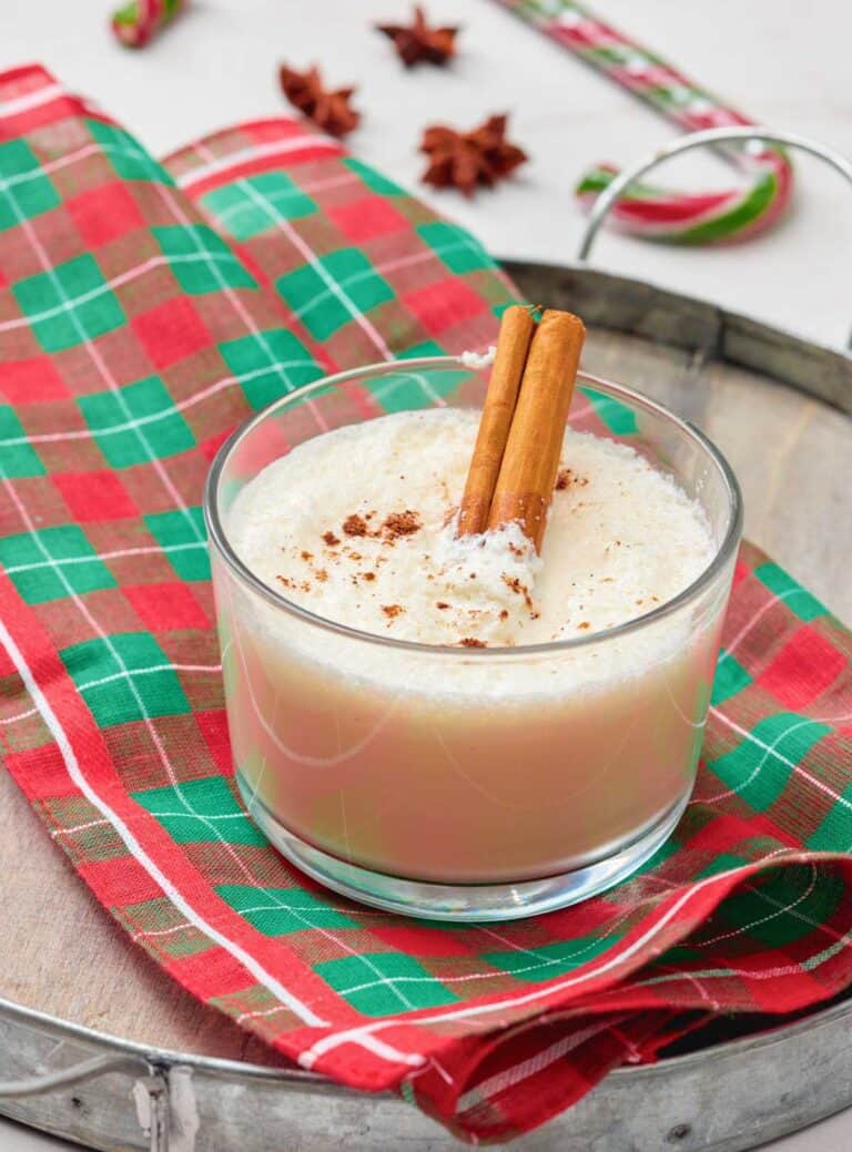 A glass of eggnog topped with ground nutmeg and two cinnamon sticks, placed on a red and green plaid napkin on a wooden tray.