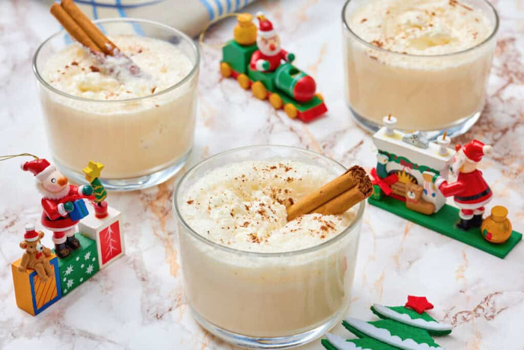 Three glasses of eggnog topped with cinnamon sticks and sprinkled spice, surrounded by small Christmas-themed figurines on a marble surface.