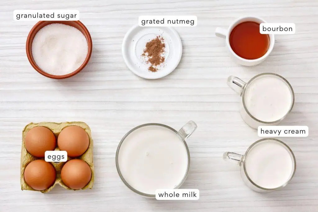 Assorted ingredients on a white surface: granulated sugar, grated nutmeg, bourbon, eggs, whole milk, and heavy cream, each in separate containers.