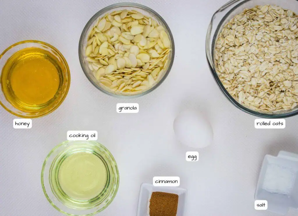Top view of separate bowls containing honey, cooking oil, granola, rolled oats, ground cinnamon, salt, and a whole egg on a white surface.