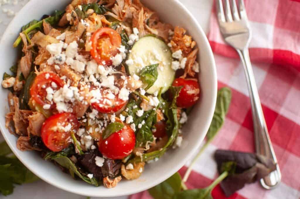 A bowl of salad with shredded meat, cherry tomatoes, spinach, cucumber slices, and crumbled cheese, placed next to a fork and a red-and-white checkered napkin.