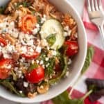 A bowl of salad with shredded meat, cherry tomatoes, spinach, cucumber slices, and crumbled cheese, placed next to a fork and a red-and-white checkered napkin.