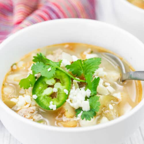 A bowl of Green Pork Posole topped with sliced jalape&ntilde;o, cilantro, and crumbled cheese, with a spoon in the bowl and a striped cloth in the background.
