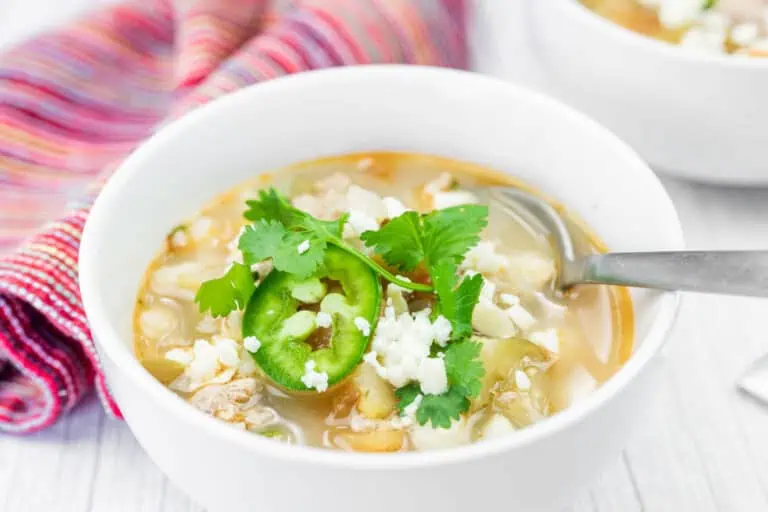 A bowl of Green Pork Posole topped with sliced jalapeño, cilantro, and crumbled cheese, with a spoon in the bowl and a striped cloth in the background.