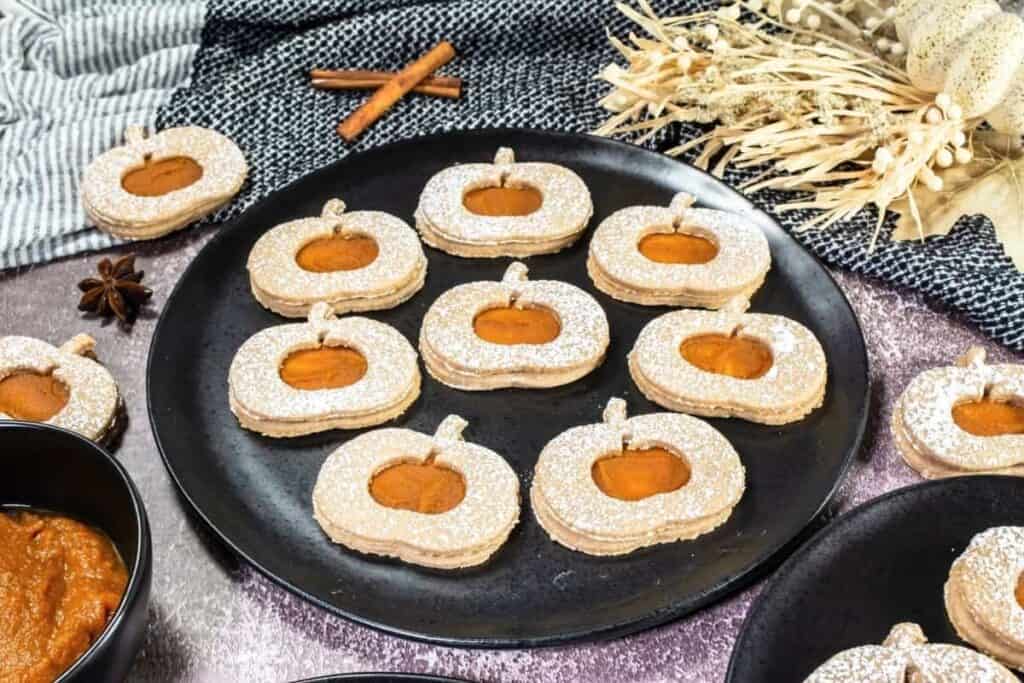A black plate with pumpkin-shaped sandwich cookies dusted with powdered sugar, filled with orange jam, set on a table with fall-themed decor.