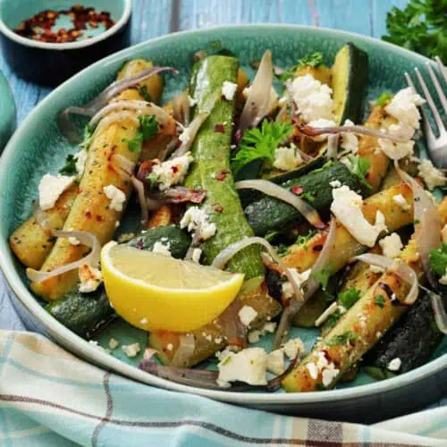 A bowl of roasted zucchini and onions topped with feta cheese, fresh herbs, and red pepper flakes, garnished with a lemon wedge; fork and napkin beside the bowl.