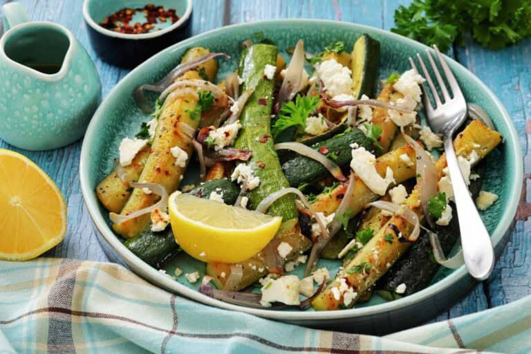 A bowl of roasted zucchini and onions topped with feta cheese, fresh herbs, and red pepper flakes, garnished with a lemon wedge; fork and napkin beside the bowl.