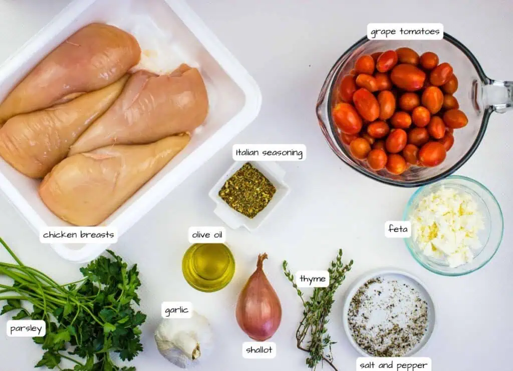 Raw chicken breasts, grape tomatoes, feta cheese, Italian seasoning, olive oil, parsley, garlic, shallot, thyme, salt, and pepper arranged on a white surface, each labeled.