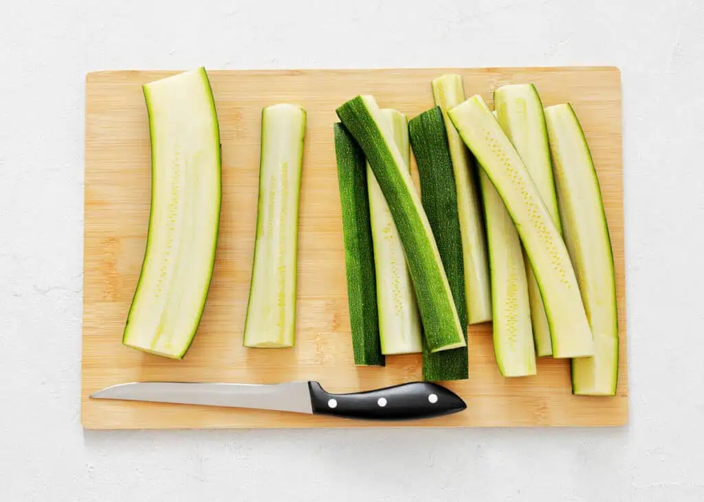 Sliced zucchini sticks and a knife are arranged on a wooden cutting board.