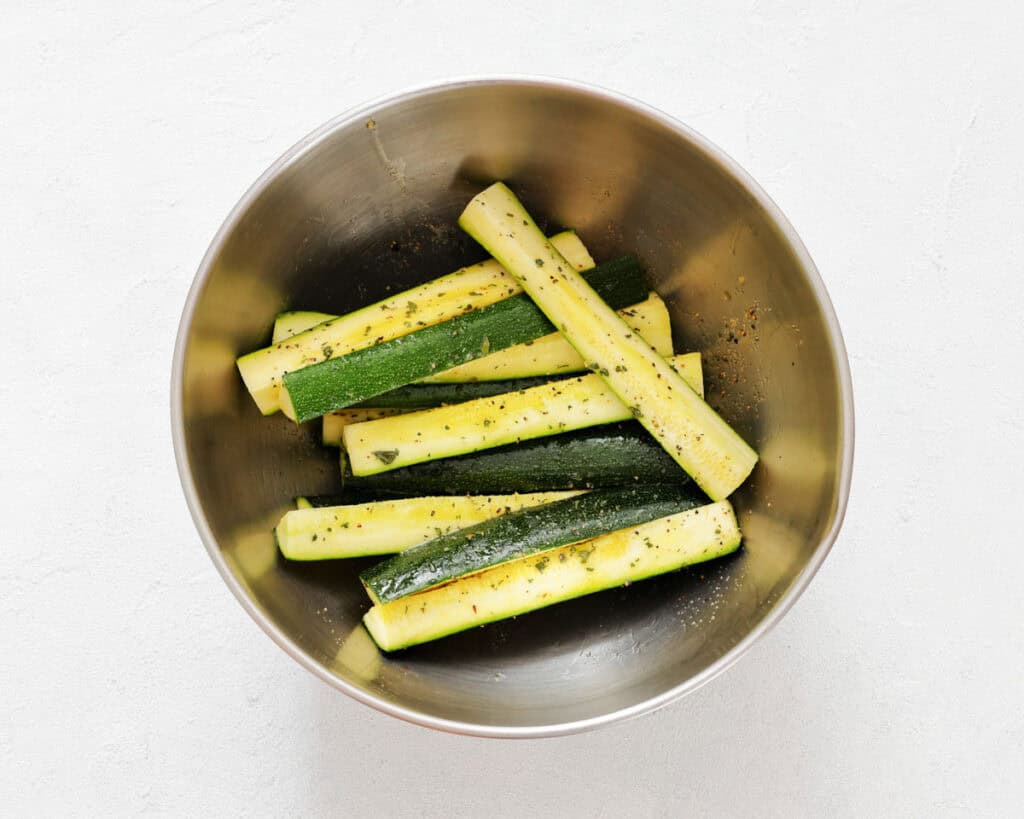 Metal bowl containing raw zucchini strips seasoned with herbs and spices, placed on a white surface.