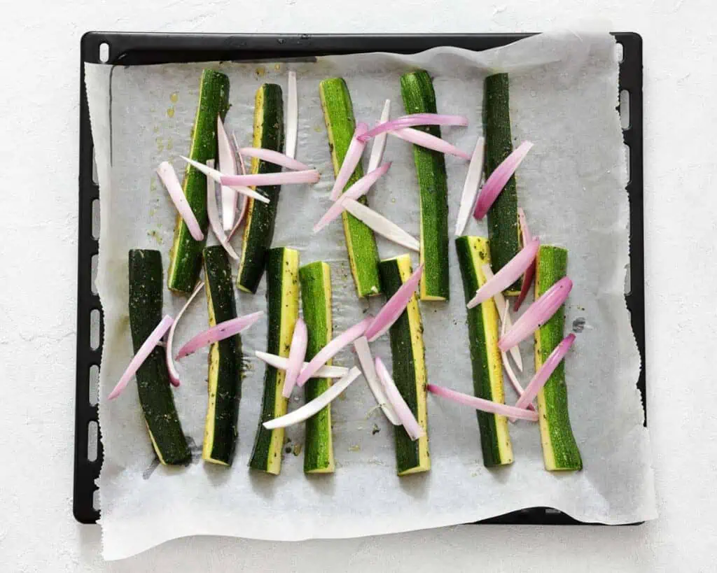 Zucchini sticks and sliced shallots arranged on a parchment-lined baking tray, ready to be roasted.