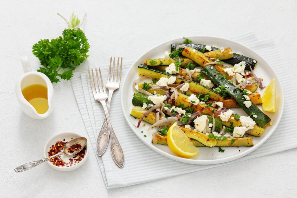 A plate of grilled zucchini with crumbled feta, herbs, and lemon wedges, served with olive oil, red pepper flakes, fresh parsley, and two forks on a striped cloth.