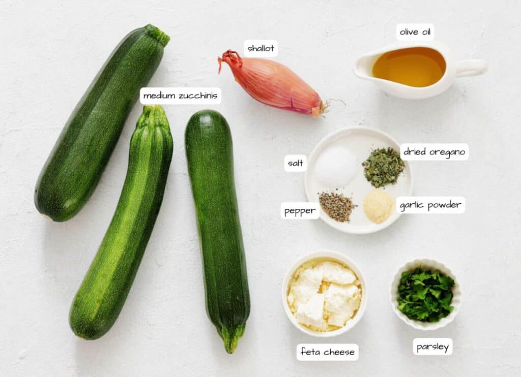 Three medium zucchinis, a shallot, olive oil, dried oregano, garlic powder, salt, pepper, feta cheese, and chopped parsley arranged on a white surface.