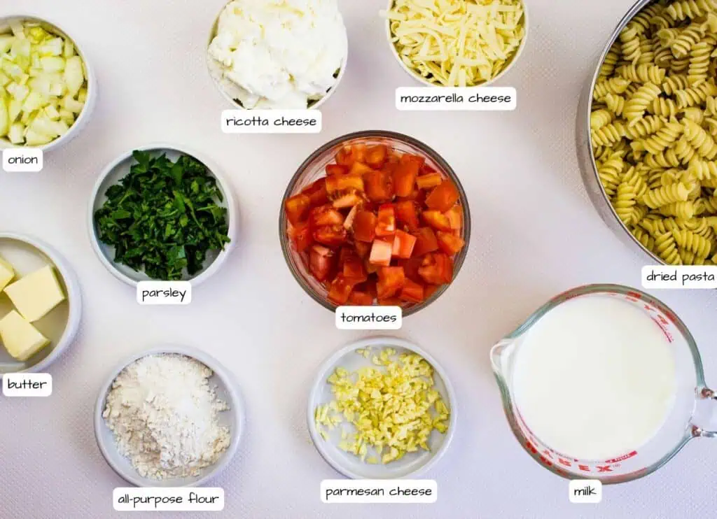 Bowls of diced onion, parsley, butter, all-purpose flour, ricotta cheese, mozzarella cheese, tomatoes, parmesan cheese, dried pasta, and a measuring cup of milk, labeled.