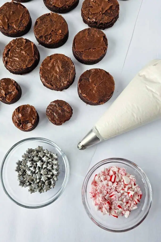 Round brownie pieces, a piping bag with white frosting, a bowl of silver star-shaped sprinkles, and a bowl of crushed peppermint on a white surface.
