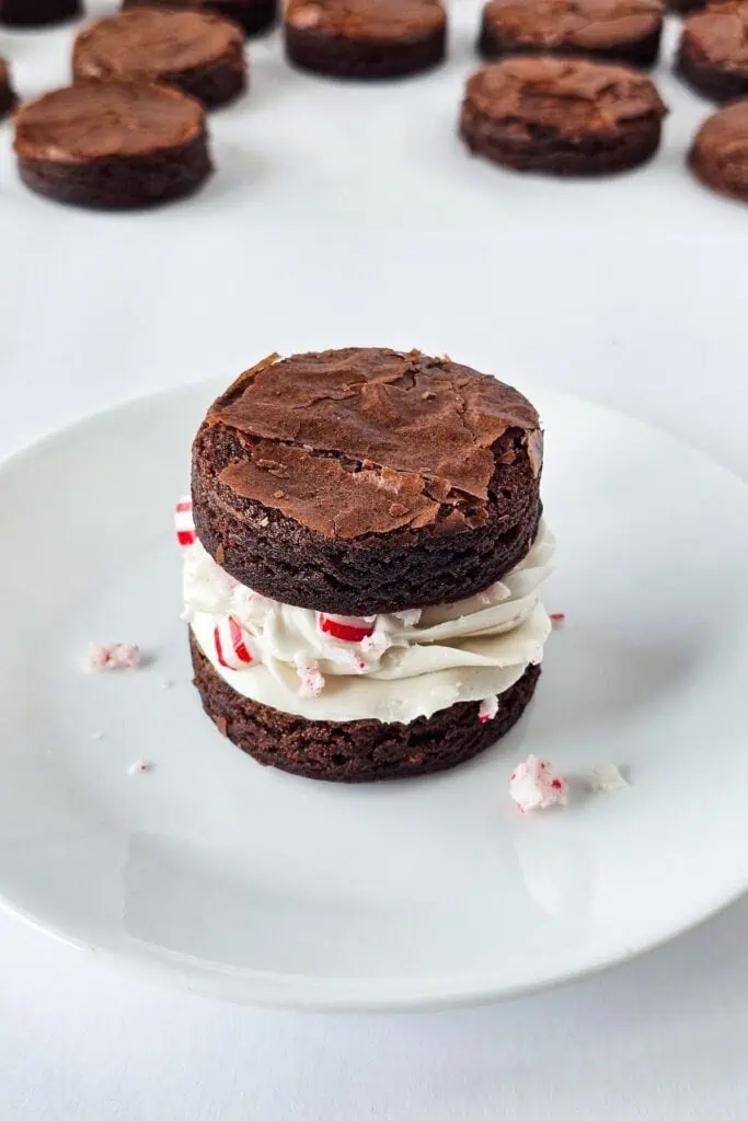 A round brownie sandwich filled with white frosting and crushed peppermint pieces sits on a white plate, with more brownie sandwiches in the background.