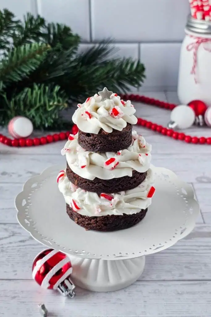 A three-tiered chocolate brownie stack with white frosting and crushed peppermint, displayed on a white cake stand with holiday decorations in the background.
