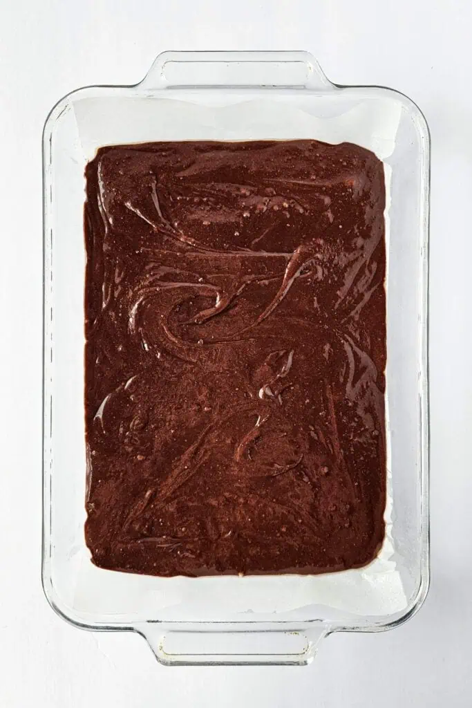 A glass baking dish lined with parchment paper is filled with an even layer of chocolate brownie batter.