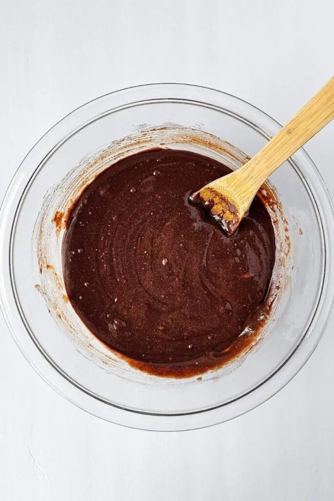 A glass bowl filled with chocolate brownie batter and a wooden spoon resting inside, set on a white surface.