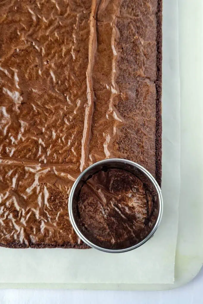 A sheet of baked brownies with a round piece cut out using a metal cookie cutter, placed on parchment paper.