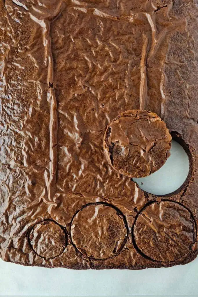 A sheet of baked brownies with several circular pieces cut out, leaving round holes and one brownie round placed near a cutout.