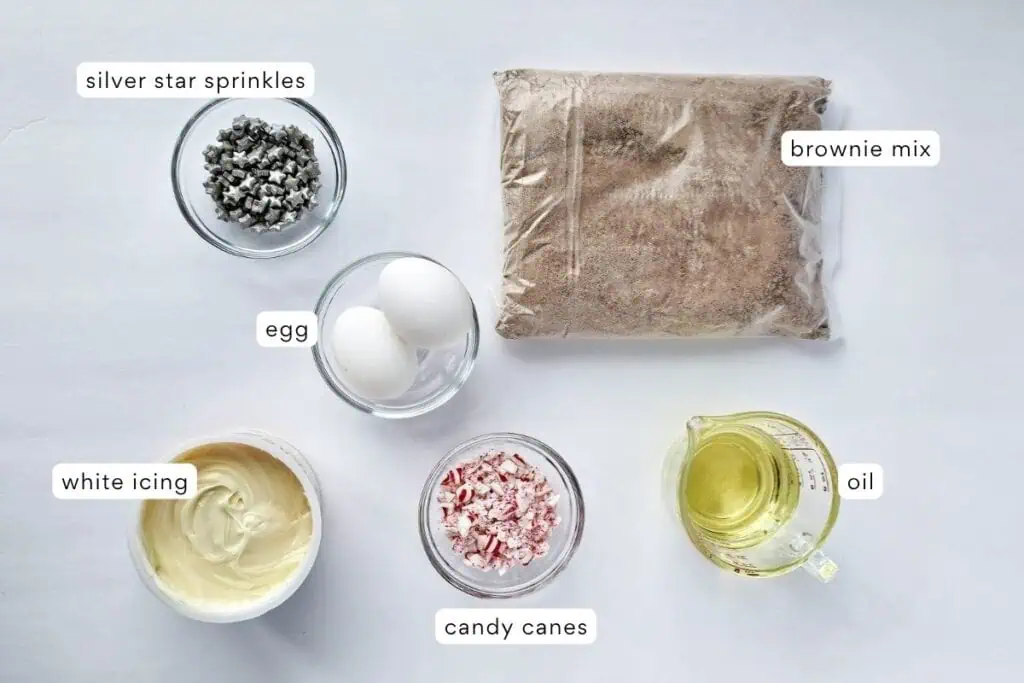 A top-down view of labeled ingredients for brownies: silver star sprinkles, brownie mix, eggs, oil, white icing, and crushed candy canes on a light surface.