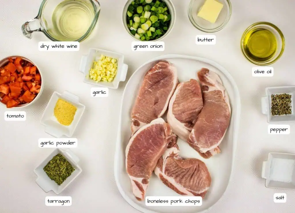 Overhead view of labeled ingredients for pork chops, including pork chops, green onion, butter, olive oil, pepper, salt, tarragon, garlic powder, garlic, tomato, and dry white wine.