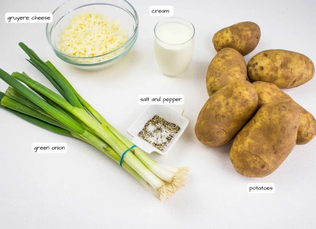 A bundle of green onions, a bowl of grated Gruyere cheese, a glass of cream, a dish of salt and pepper, and several russet potatoes on a white surface.