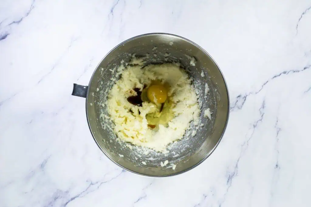A metal mixing bowl containing creamed butter and sugar with an egg and vanilla extract on top, ready to be mixed further, on a white marble surface.
