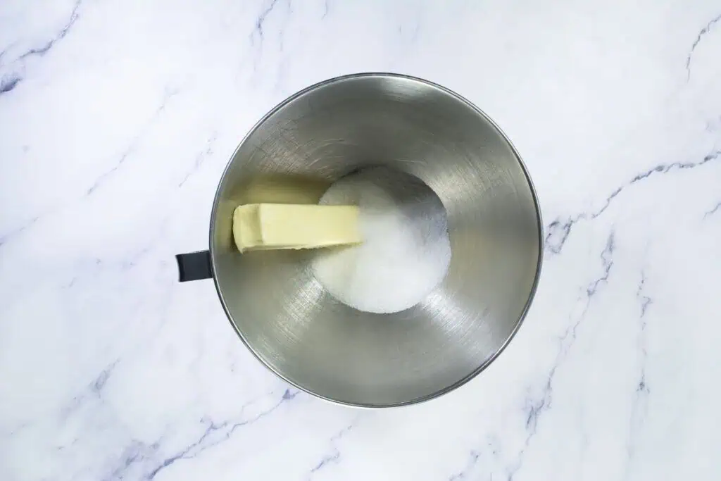 A stick of butter and a pile of granulated sugar in a stainless steel mixing bowl on a marble countertop.