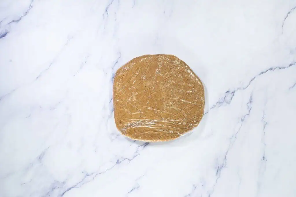 A round sheet of dough wrapped in plastic wrap sits on a white marble surface.