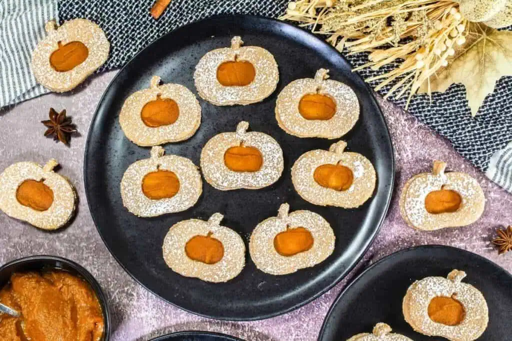 A black plate holds pumpkin-shaped cookies dusted with powdered sugar, each with pumpkin filling. More cookies and pumpkin puree are arranged nearby on a textured surface.