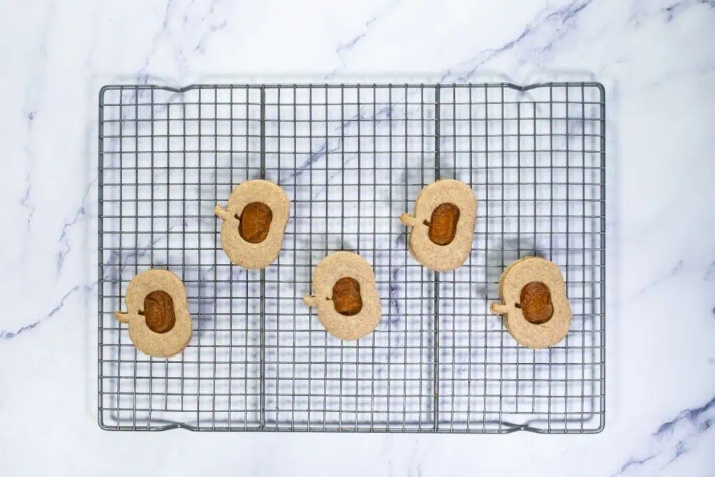 Five pear-shaped cookies with cut-out centers are arranged on a metal cooling rack over a white marble surface.