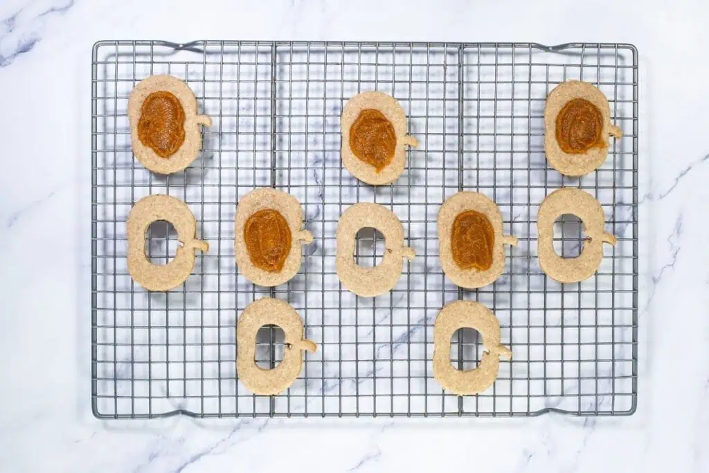 Ten oval-shaped cookies with pumpkin stems, some topped with brown filling and others with cut-out centers, are arranged on a wire cooling rack over a marble surface.