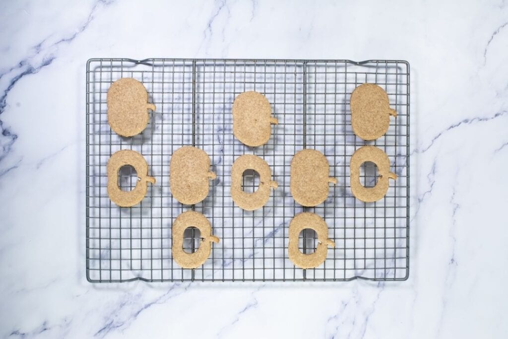 Ten oval-shaped cookies with small notches are arranged on a cooling rack placed on a marble surface. Four cookies have a square hole in the center.