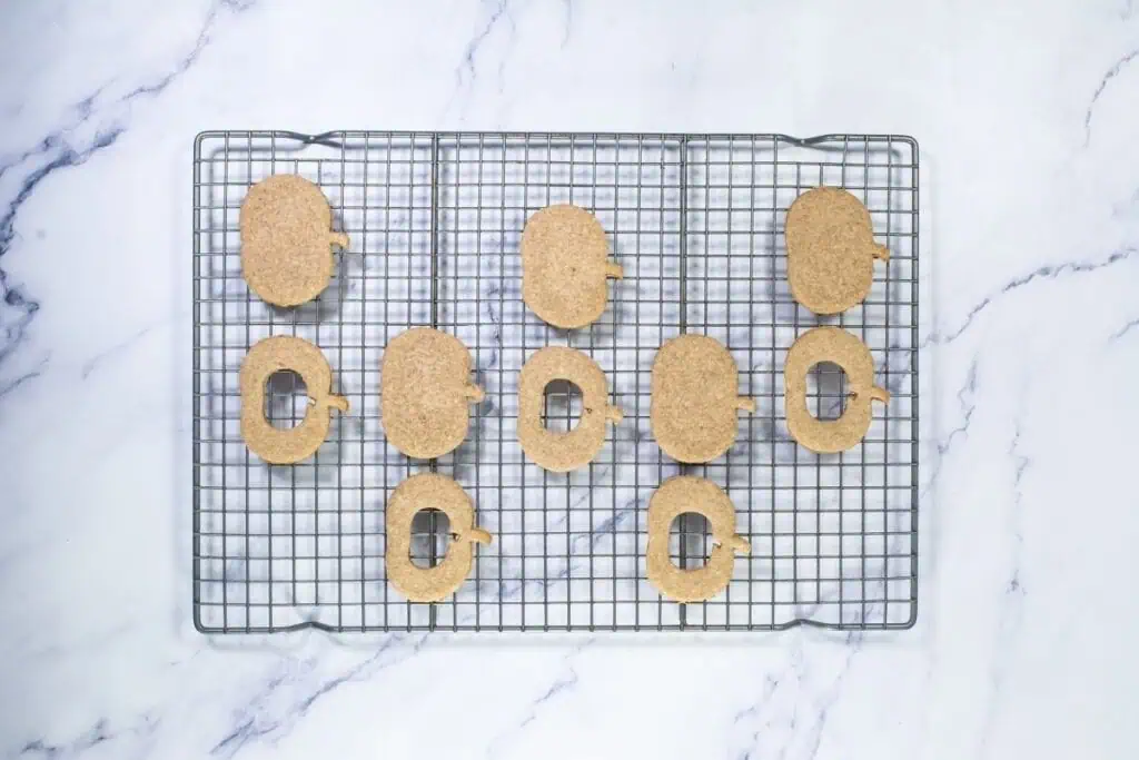 Ten oval-shaped cookies with small notches are arranged on a cooling rack placed on a marble surface. Four cookies have a square hole in the center.