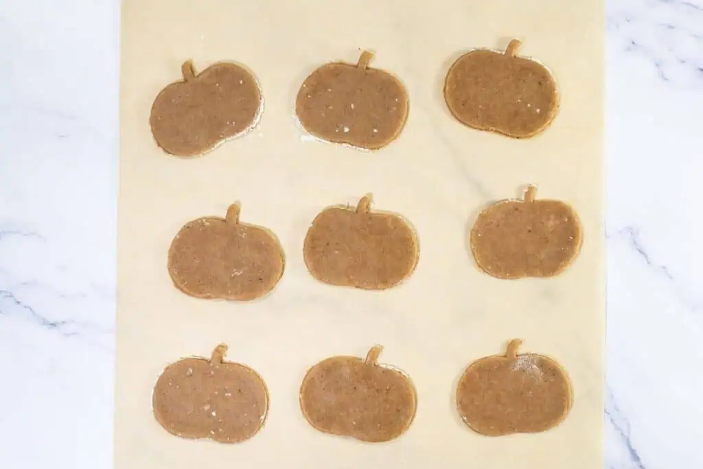 Nine pumpkin-shaped cookie dough cutouts are arranged in rows on a parchment-lined baking sheet.