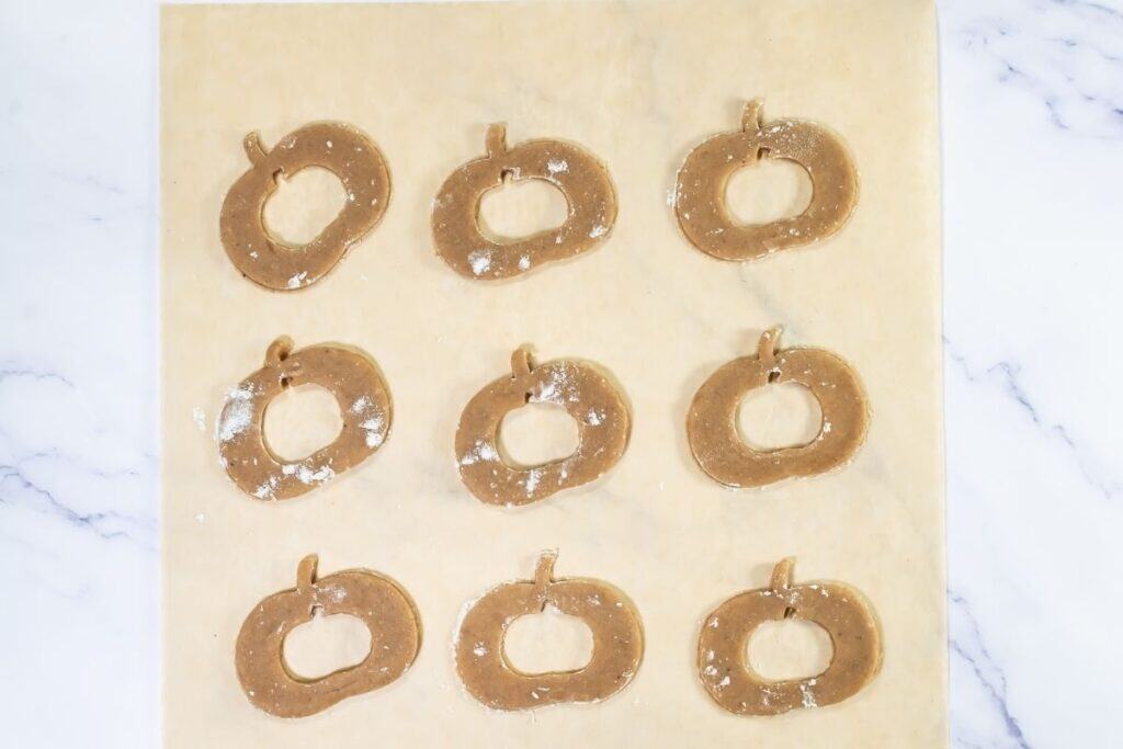 Nine pumpkin-shaped cookie dough pieces are arranged in three rows on a sheet of parchment paper, lightly dusted with flour, ready for baking.