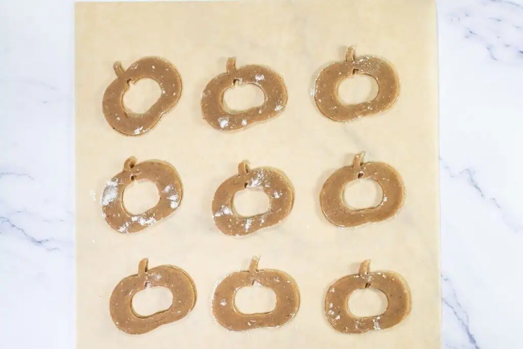 Nine pumpkin-shaped cookie dough pieces are arranged in three rows on a sheet of parchment paper, lightly dusted with flour, ready for baking.