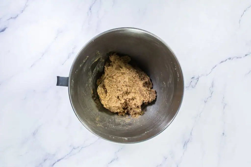 A metal mixing bowl with a thick dough mixture sits on a white marble surface.