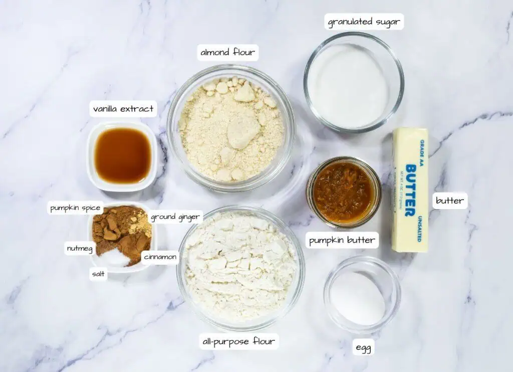 Top-down view of labeled baking ingredients in bowls and containers on a white surface, including flours, sugar, spices, pumpkin butter, vanilla extract, butter, and an egg.