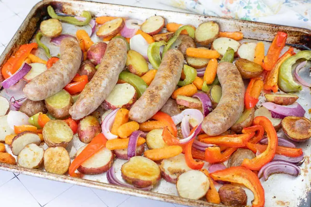 A sheet pan filled with cooked sausages, sliced potatoes, carrots, red and green bell peppers, and red onions.
