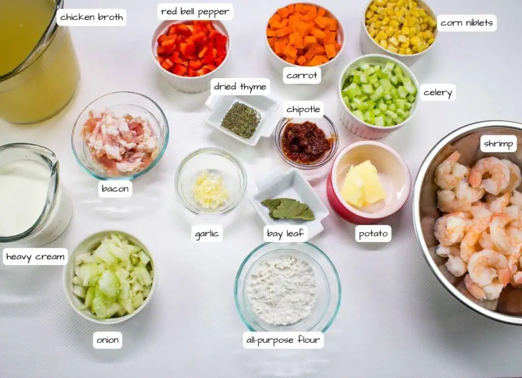 An assortment of labeled ingredients in bowls and containers, including shrimp, vegetables, chicken broth, bacon, heavy cream, spices, and flour, arranged on a white surface.