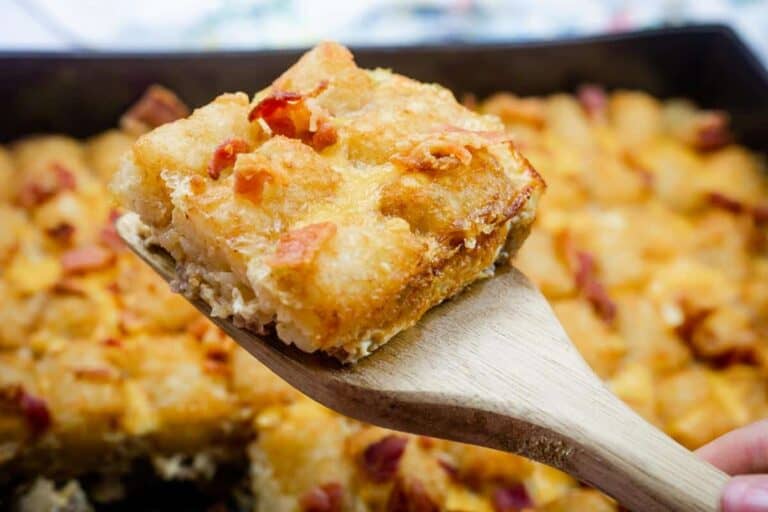 A wooden spatula holds a square slice of cheesy breakfast casserole with bacon on top, with the rest of the casserole in the background.