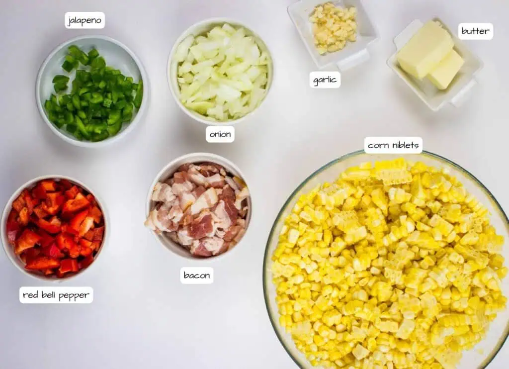 Top view of chopped jalapeno, onion, red bell pepper, bacon, corn niblets, garlic, and butter in separate bowls on a white surface, each labeled.