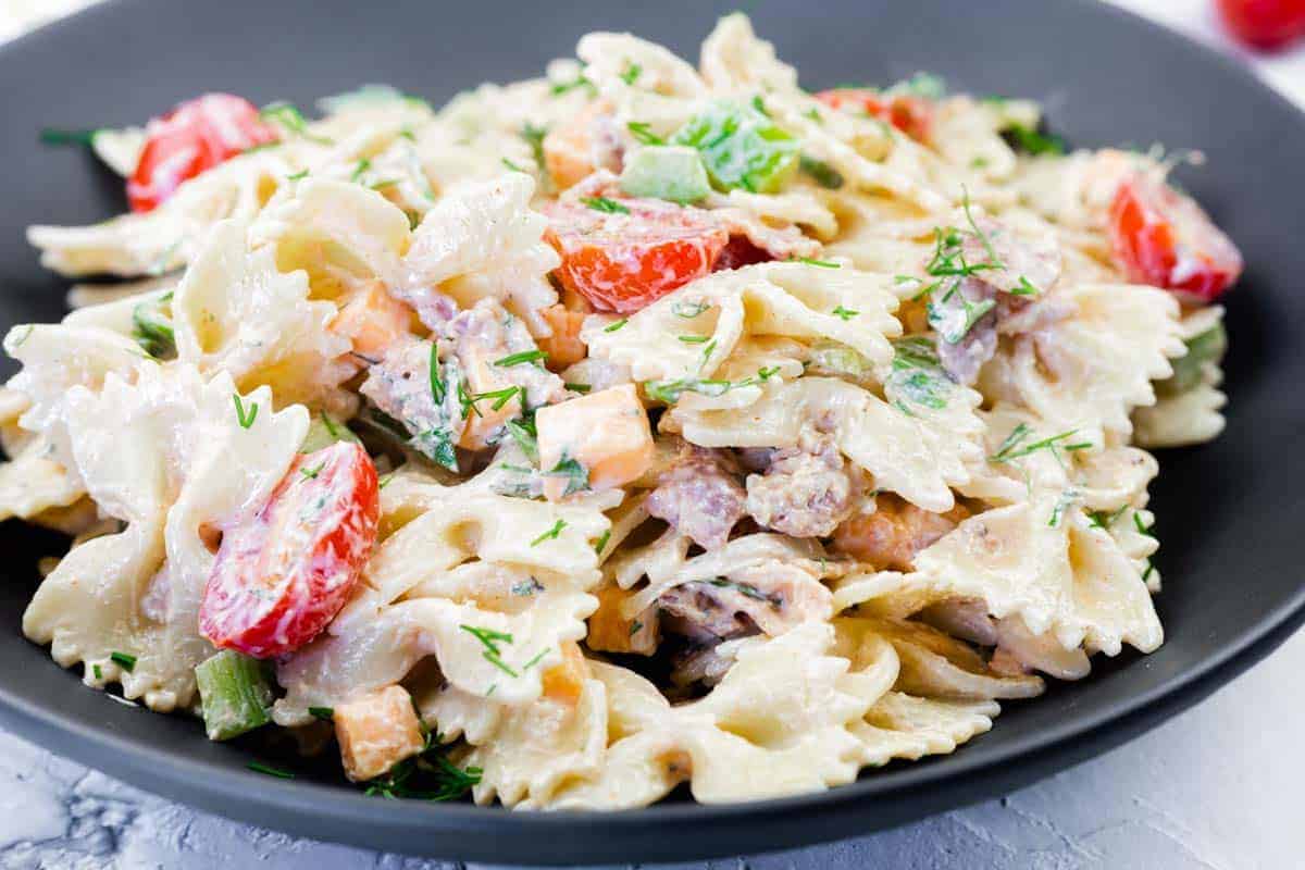 A black plate filled with creamy bowtie pasta, cherry tomatoes, diced vegetables, and herbs.