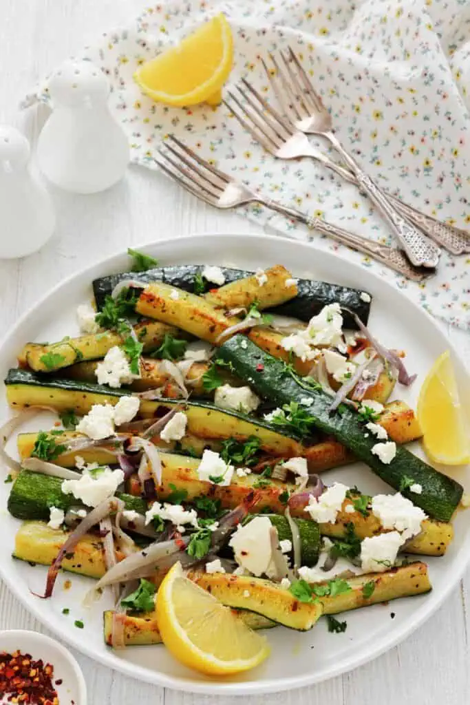 A plate of grilled zucchini topped with crumbled feta cheese, sliced red onions, chopped herbs, and lemon wedges, with forks and a napkin in the background.