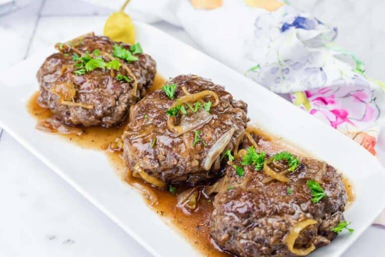 Three cooked Salisbury steaks topped with onions and brown gravy, garnished with parsley, served on a rectangular white plate.