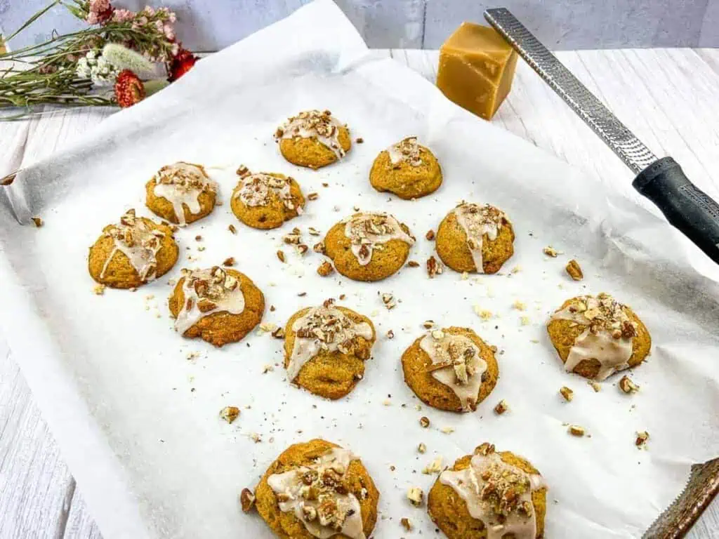 A baking tray with twelve frosted cookies topped with chopped nuts, placed on parchment paper beside a grater, a block of brown sugar, and a small bouquet of flowers.