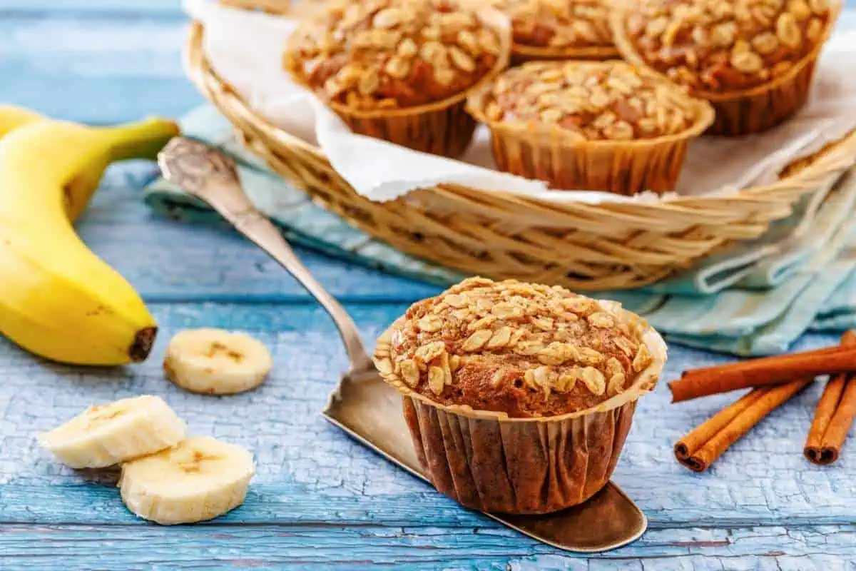 Banana oatmeal muffins on a metal spatula and in a basket, with sliced bananas and cinnamon sticks on a blue wooden surface.